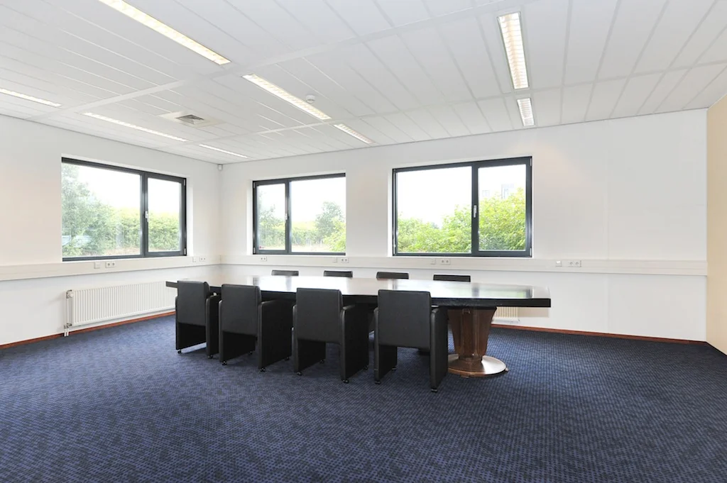 Vergaderruimte aan de Laan van S'Gravenmade met groot donkerhouten tafel en acht zwarte stoelen, omgeven door ramen met uitzicht op groen.